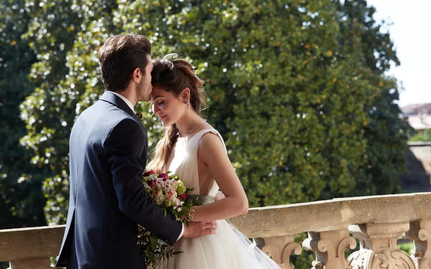 Newlywed couple sharing a tender moment, with the groom kissing the bride’s forehead, as she holds a bouquet of flowers.
