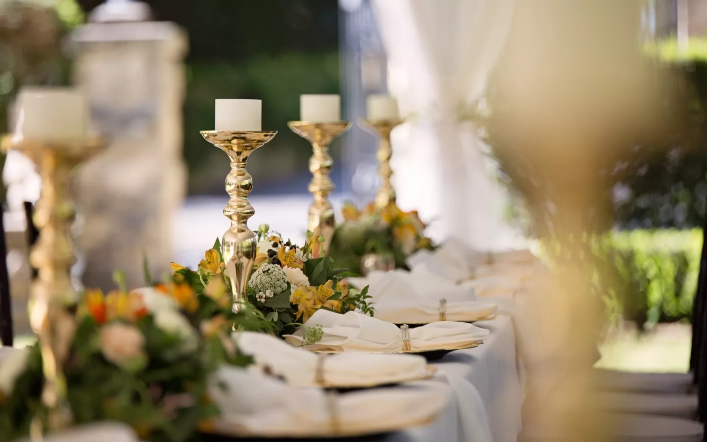 Wedding reception table with gold candleholders, white plates, and floral arrangements of yellow, orange, and white flowers.